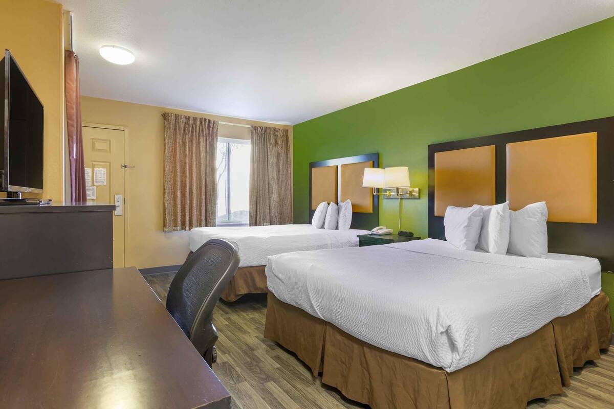 A hotel room with two beds, a desk, a television, and a window with curtains.