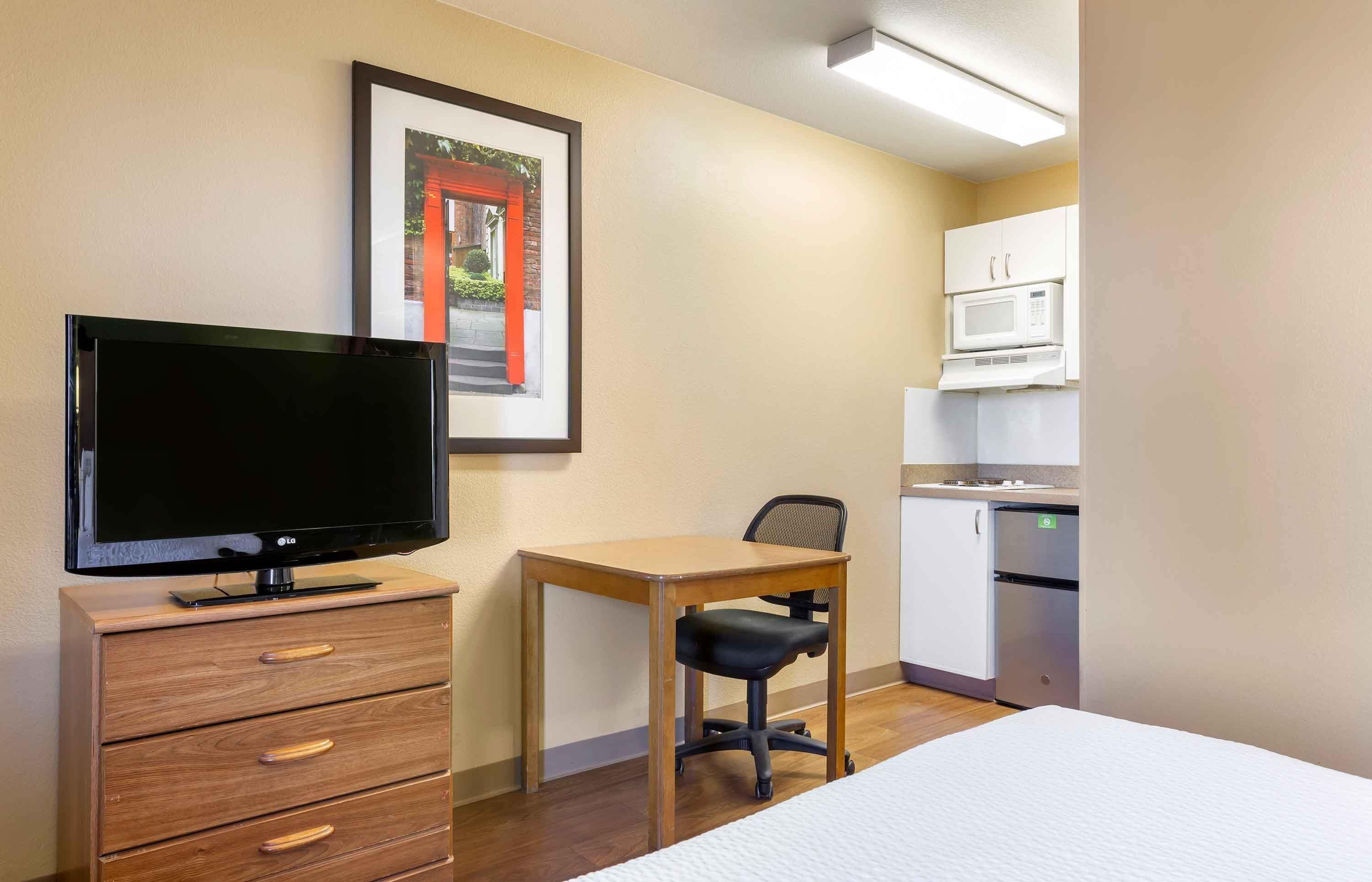 Une chambre d’hôtel équipée d’une télévision, d’une commode en bois, d’un bureau, d’une chaise, d’un lit, d’une kitchenette avec micro-ondes et de placards.