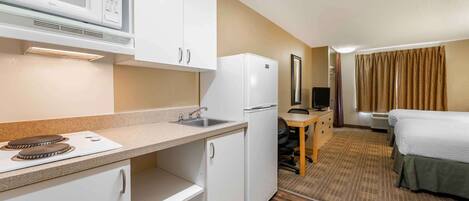 Een compacte hotelkamer met een kitchenette, inclusief een magnetron, koelkast en kookplaat. Er is een bed met een groene sprei en een klein bureau met een stoel.