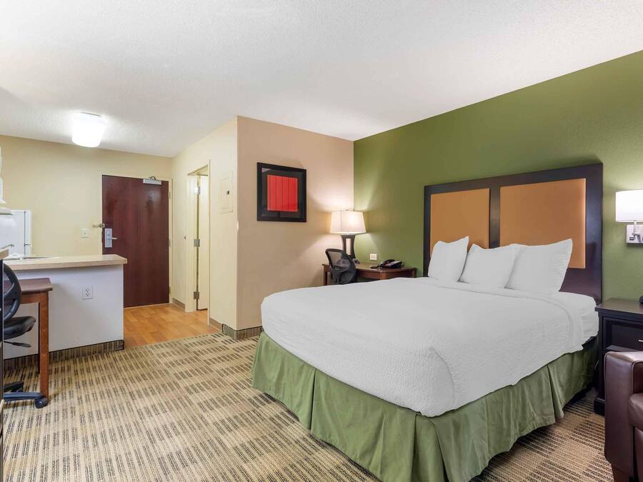 Quarto de hotel com cama, escrivaninha, cadeira e um pequeno espaço de cozinha.