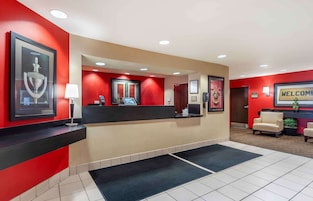 Uma área de recepção de hotel com esquema de cores vermelho e bege, um balcão de recepção, um troféu emoldurado, uma placa de 'Welcome' e duas poltronas.