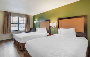 Habitación de hotel con dos camas, una ventana con cortinas, una silla y dos lámparas fijadas a la pared.