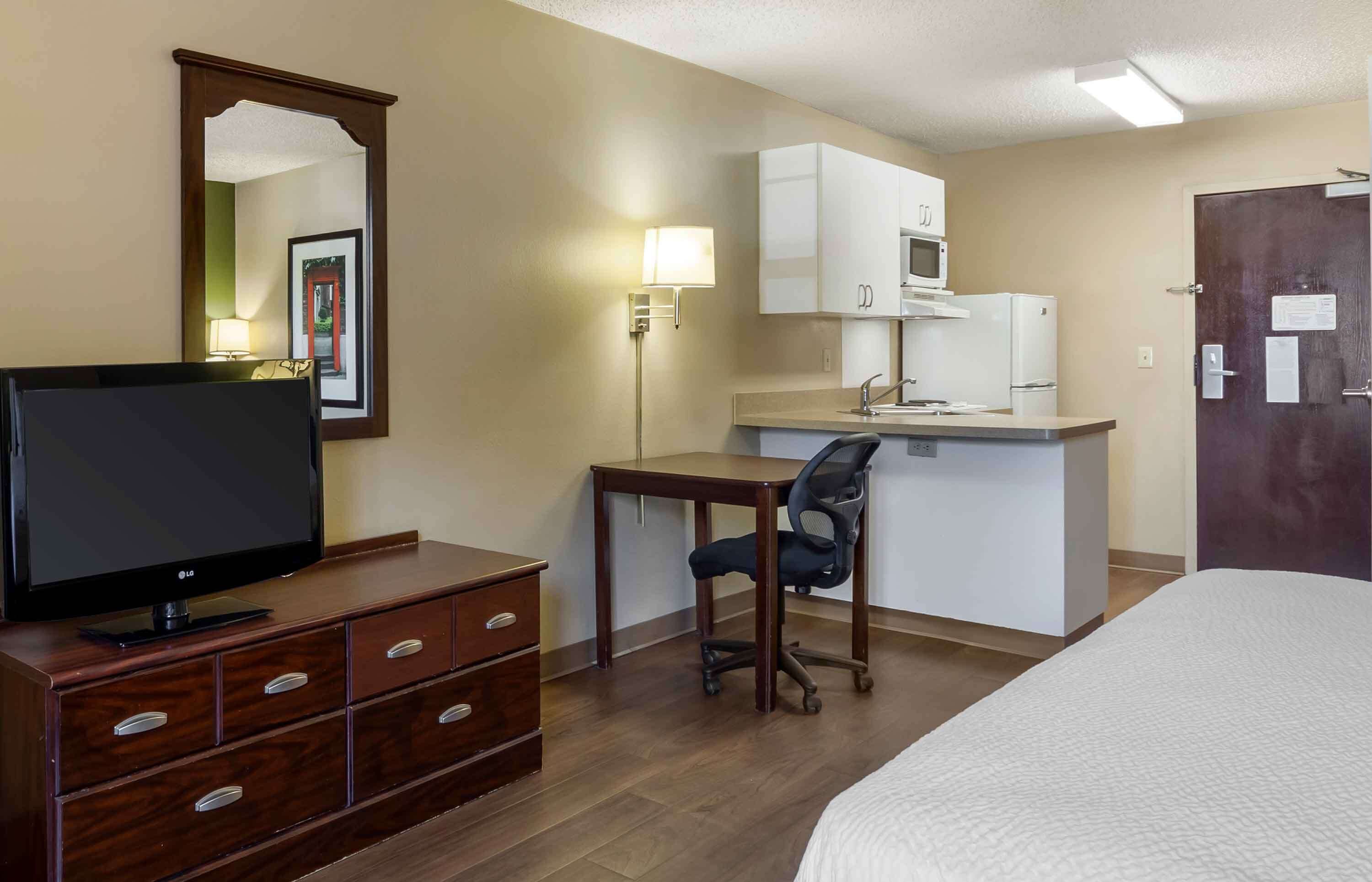Habitación de hotel con cama, televisor en una cómoda de madera, escritorio con silla, kitchenette y una puerta.