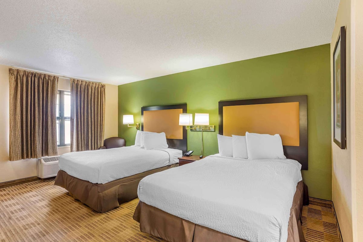 A hotel room with two beds, a large window with curtains, a wall-mounted air conditioner, and a framed picture on the wall.