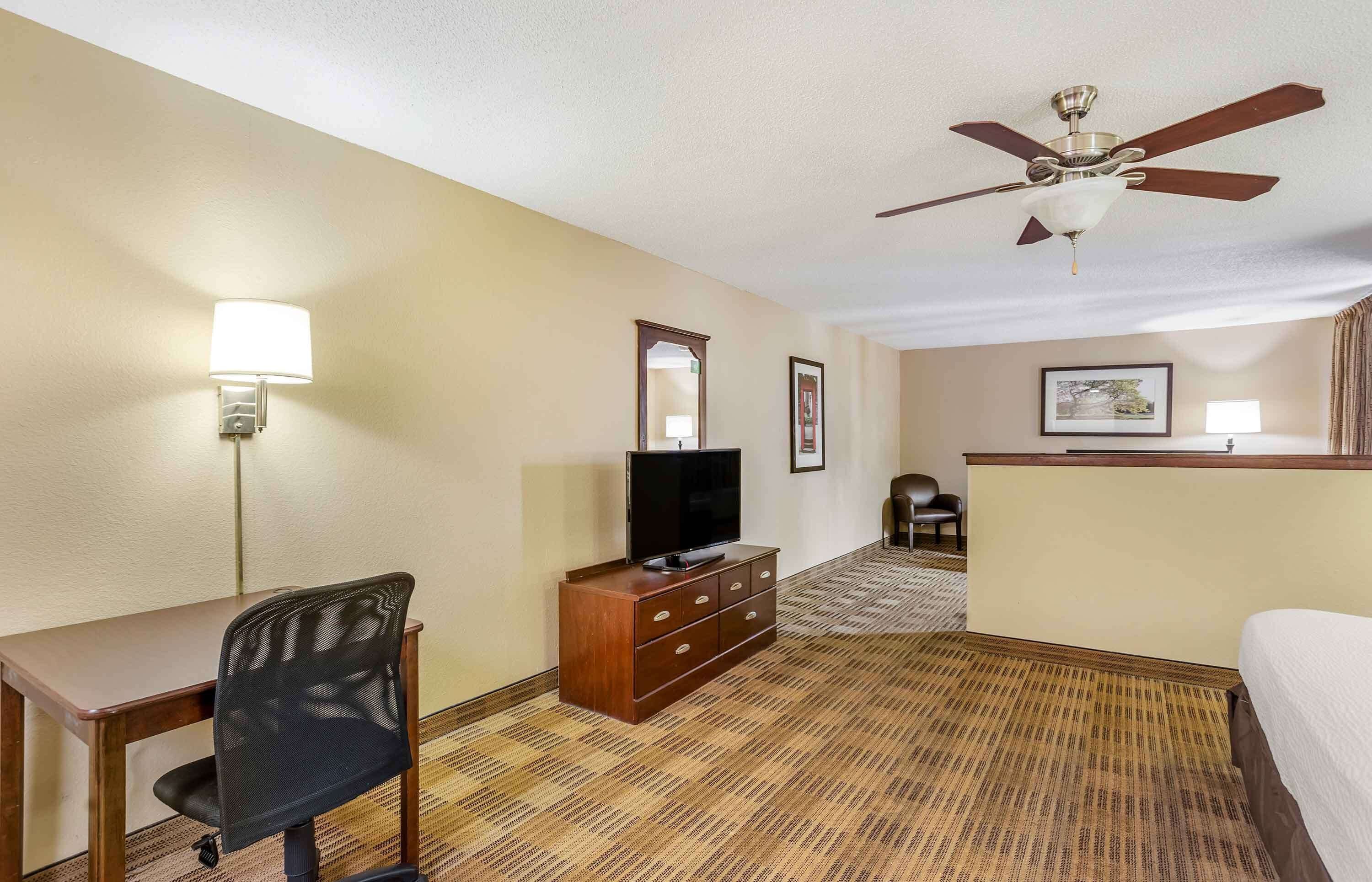 A hotel room with a bed, a desk with a chair, a television on a wooden dresser, a ceiling fan, and a carpeted floor.