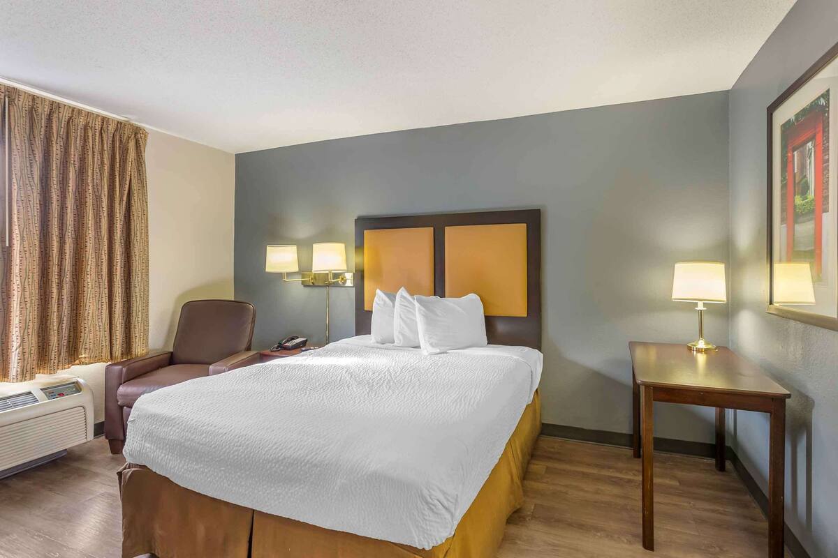 A hotel room with a large bed, two bedside lamps, a chair, a desk, and a framed picture on the wall.