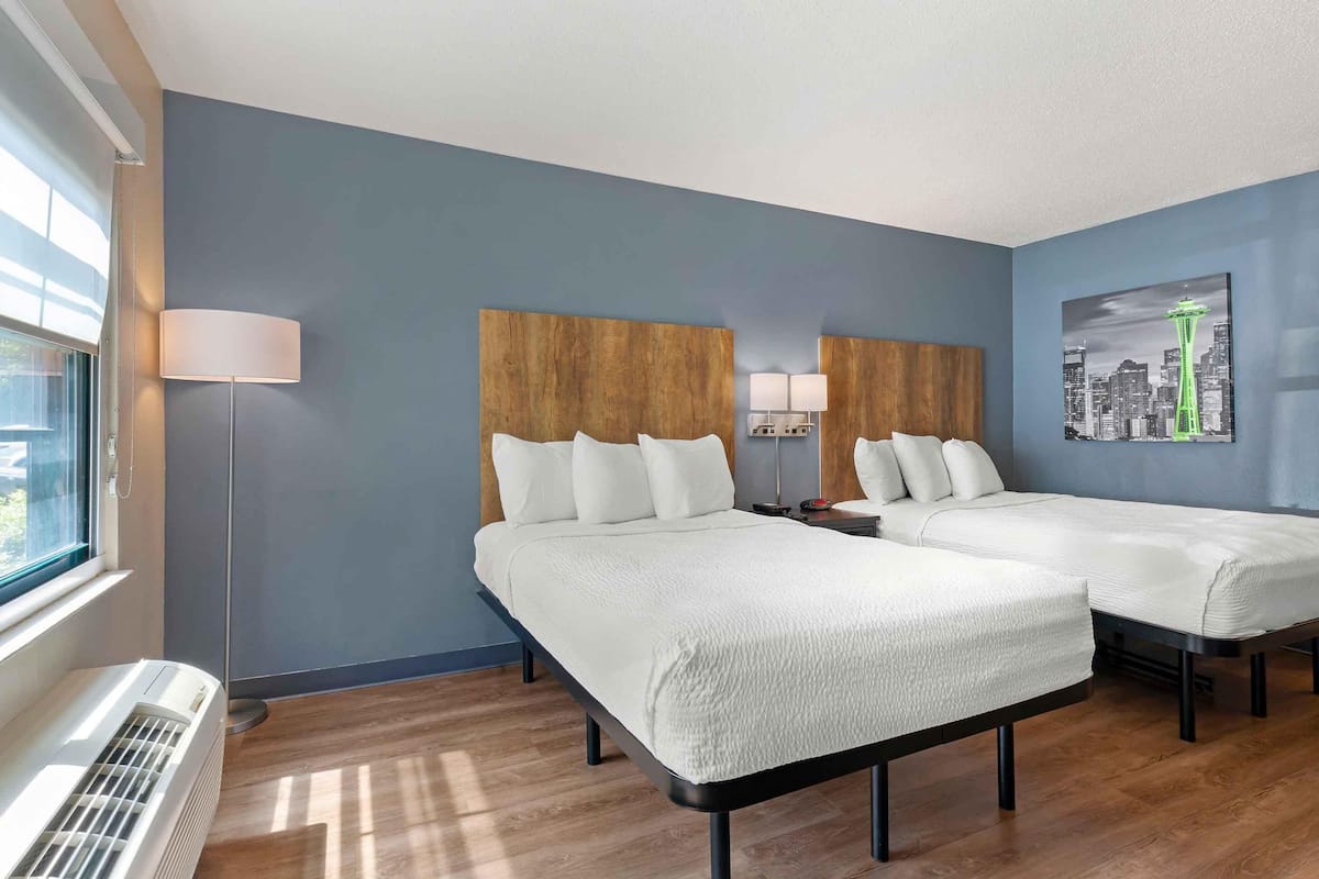 Een hotelkamer met twee bedden, een groot raam, een staande lamp en een stadsgezicht schilderij.