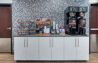 A self-service beverage station with multiple coffee dispensers, a variety of snack options, and a decorative wall with a textured pattern.