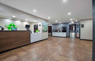 A hotel reception area with a wooden reception desk, a digital kiosk, a large green logo, a map, and a coffee machine.