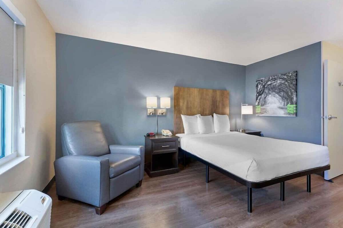 Een slaapkamer met een bed, een stoel, een nachtkastje, een wandlamp en een schilderij.