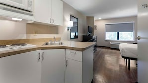 Cocina moderna con armarios blancos, microondas, estufa y lavaplatos. Hay una ventana con vistas a árboles y una cama en la habitación contigua.