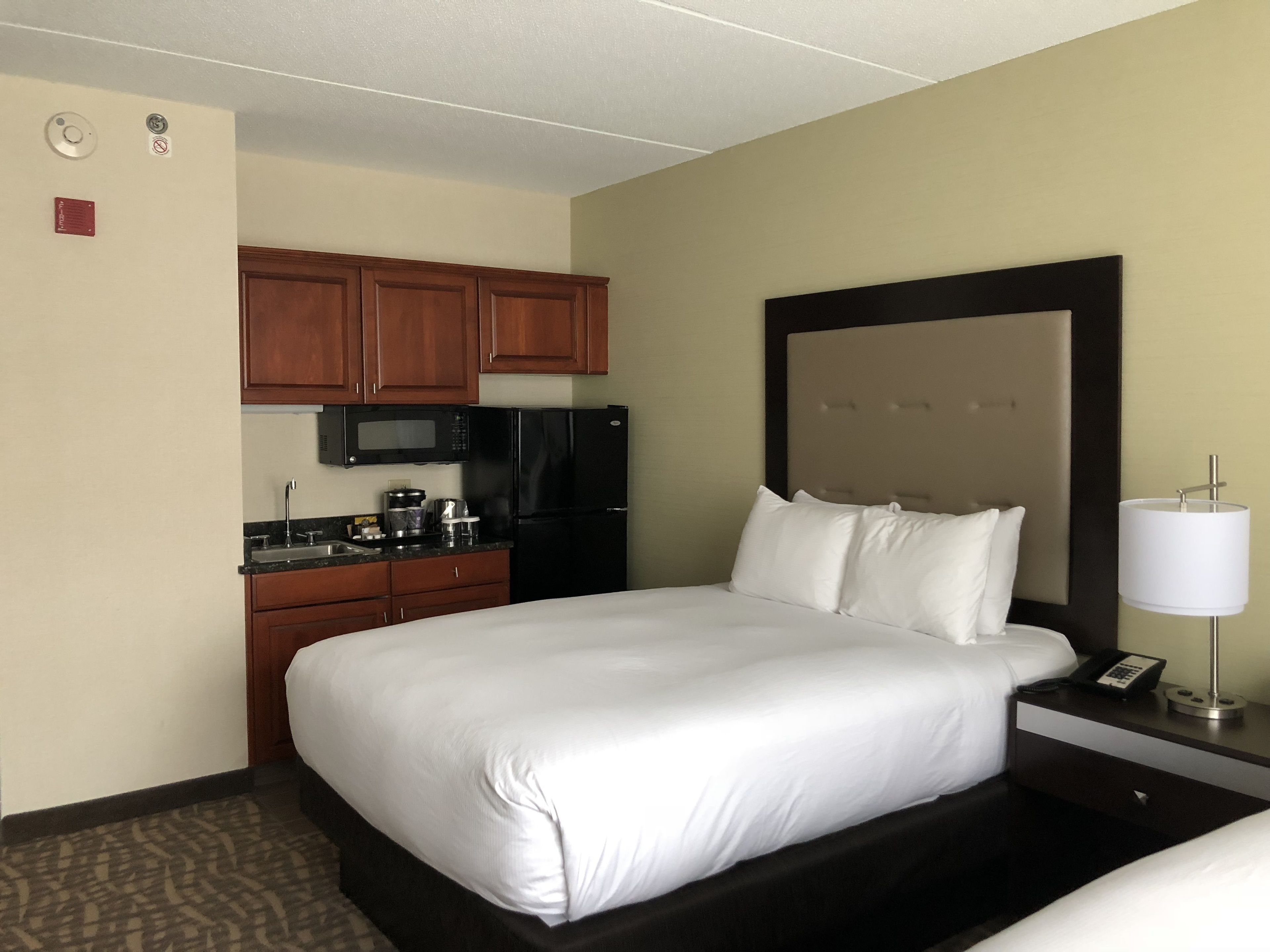 A hotel room with a bed, a small kitchenette, a microwave, a refrigerator, and a bedside table with a lamp.