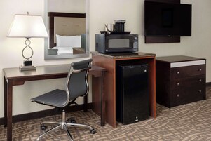 A hotel room with a desk, chair, microwave, refrigerator, TV, and a mirror.