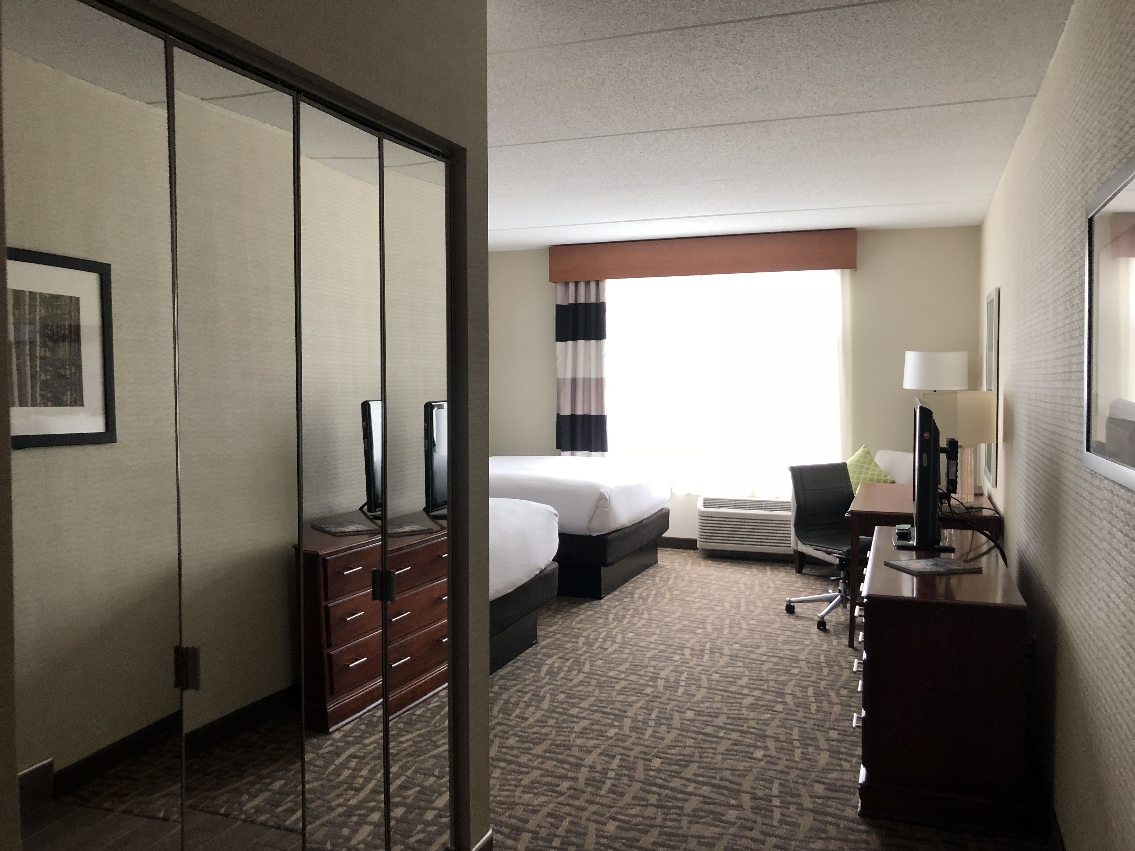 A hotel room with a bed, a desk with a lamp, a chair, a dresser, and a large window with curtains.