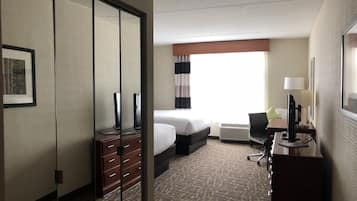 A hotel room with a bed, a desk with a lamp, a chair, a dresser, and a large window with curtains.