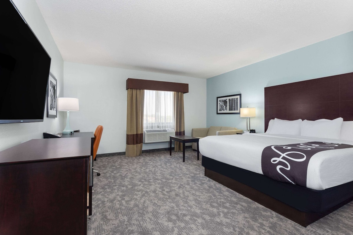 Ein Hotelzimmer mit einem großen Bett, einem Schreibtisch, einem Sessel, einem Fernseher und einem Fenster mit Vorhängen.