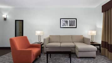 A hotel room with a sectional sofa, an armchair, two bedside tables with lamps, a framed picture on the wall, and a door.