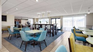 Un espace de restauration moderne avec plusieurs tables et chaises, une cuisine à l’arrière-plan et de grandes fenêtres laissant entrer la lumière naturelle.