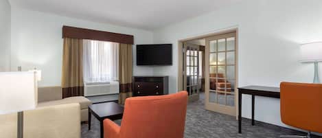 Ein Hotelzimmer mit Sofa, Sessel, Schreibtisch, Fernseher und Blick durch eine Tür nach draußen.