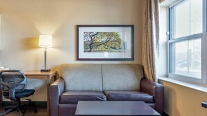 A hotel room with a sofa, a small table, a desk with a chair, a lamp, and a framed picture on the wall.
