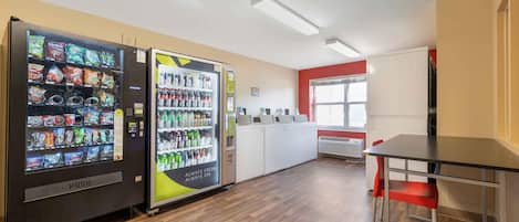 A clean, modern kitchen area with a vending machine stocked with snacks and drinks, a white refrigerator, and a red accent wall.
