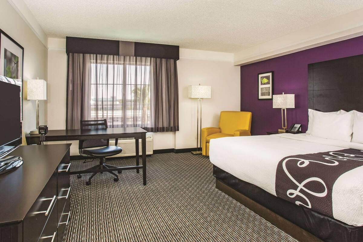 Habitación de hotel con cama, escritorio, silla, lámpara y ventana con cortinas.