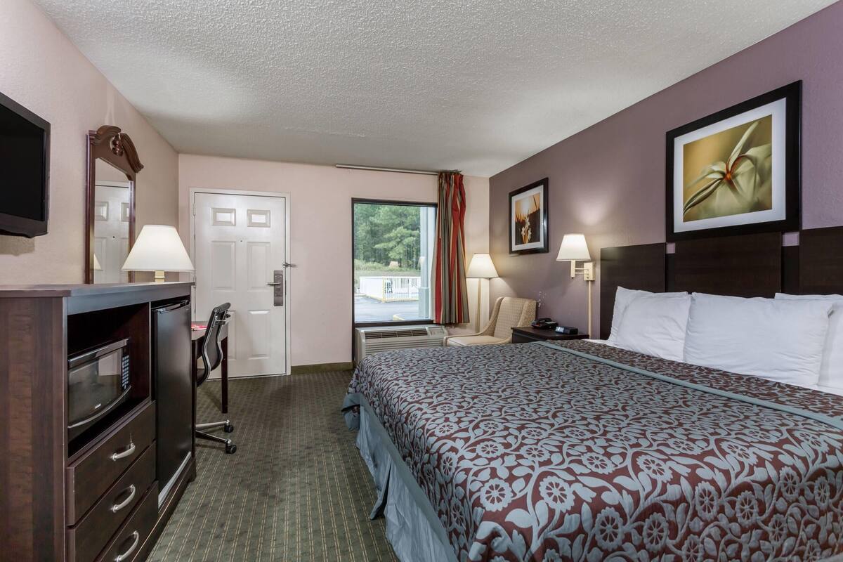 Ein Hotelzimmer mit Bett, Schreibtisch, Stuhl, Fernseher, Fenster mit Vorhängen und einer Tür.