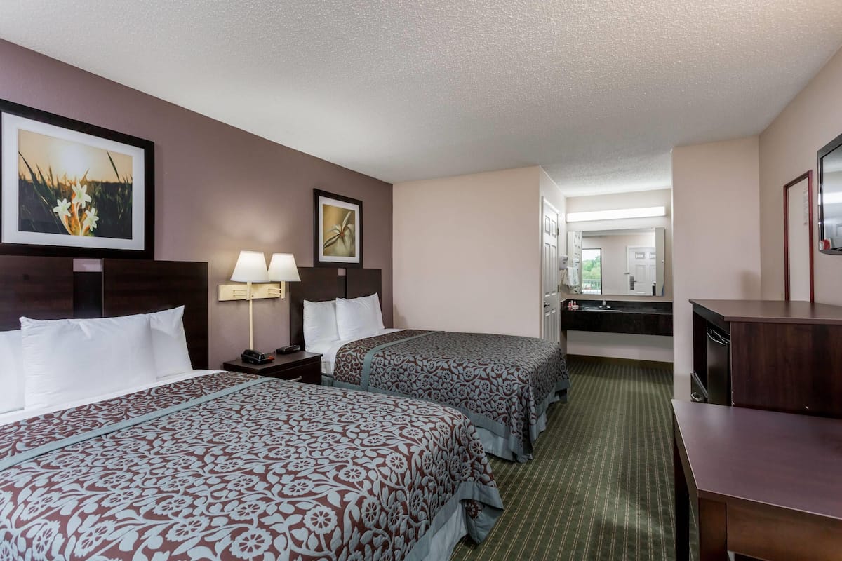 Ein Hotelzimmer mit zwei Betten, einem Schreibtisch, einem Spiegel und einem im Hintergrund sichtbaren Badezimmer.