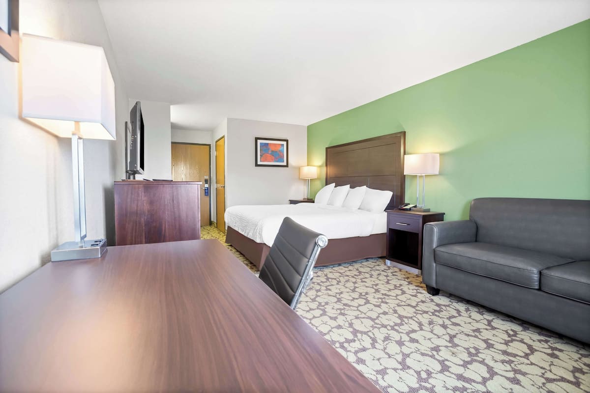 A hotel room with a wooden desk, a bed with white linens, a grey sofa, and a patterned carpet.
