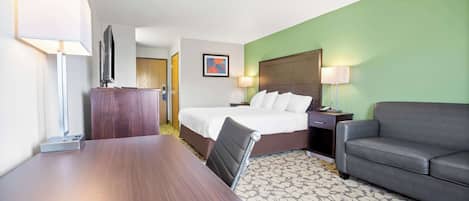 A hotel room with a wooden desk, a bed with white linens, a grey sofa, and a patterned carpet.