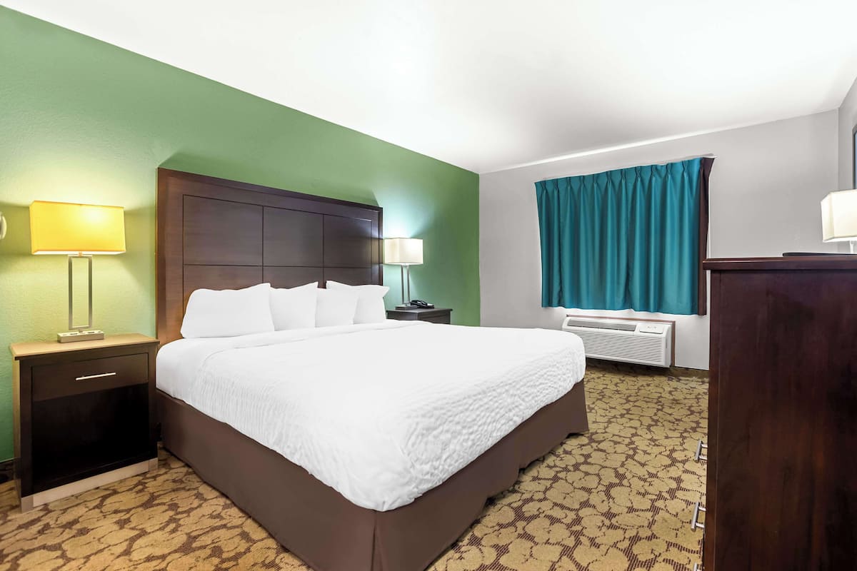A hotel room with a large bed, a nightstand with a lamp, a dresser, and a window with curtains.