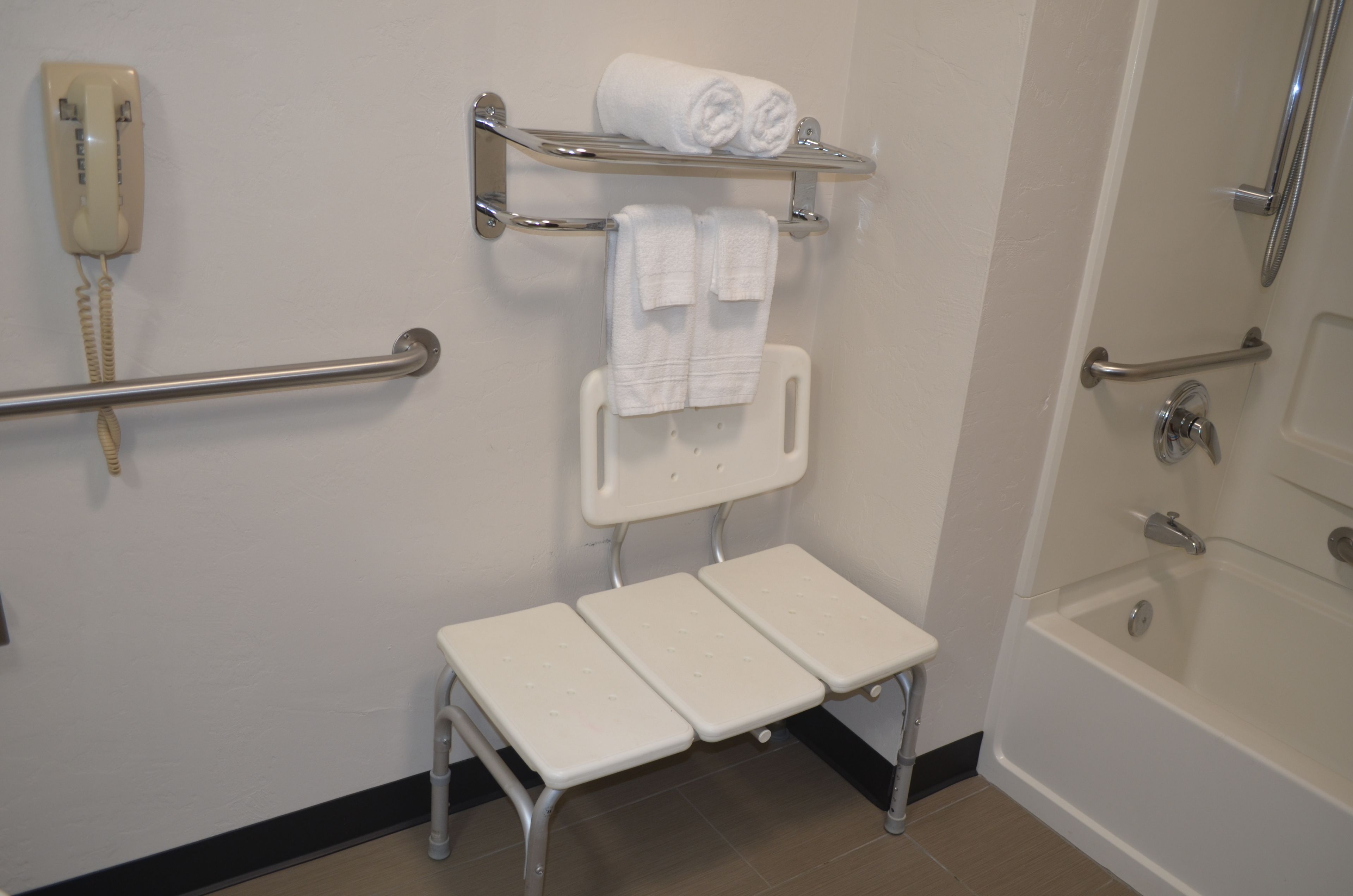 Ein Badezimmer mit Toilette, Waschbecken, Dusche und einer Bank – alles in Weiß. An der Wand befindet sich ein Handtuchhalter mit zwei Handtüchern und ein Telefon.