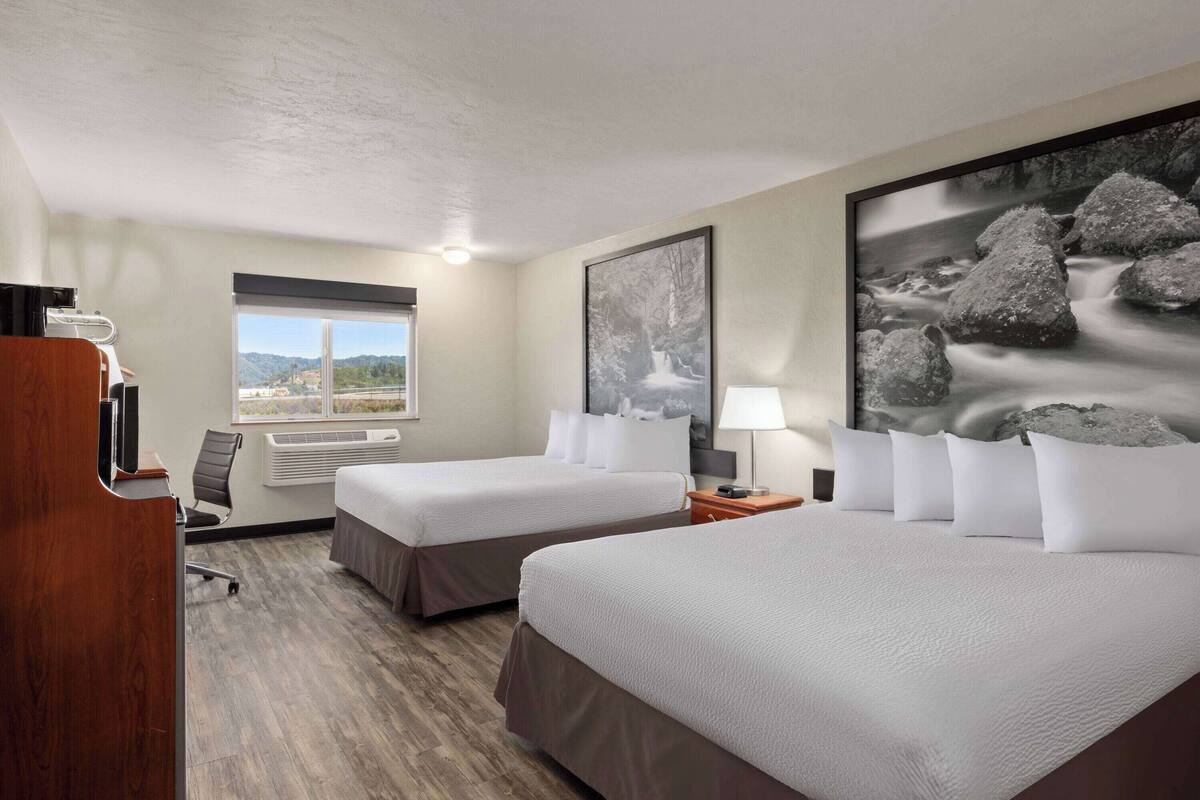 Une chambre d’hôtel avec deux lits, un bureau, une chaise, une fenêtre donnant sur un beau paysage et un tableau au mur.