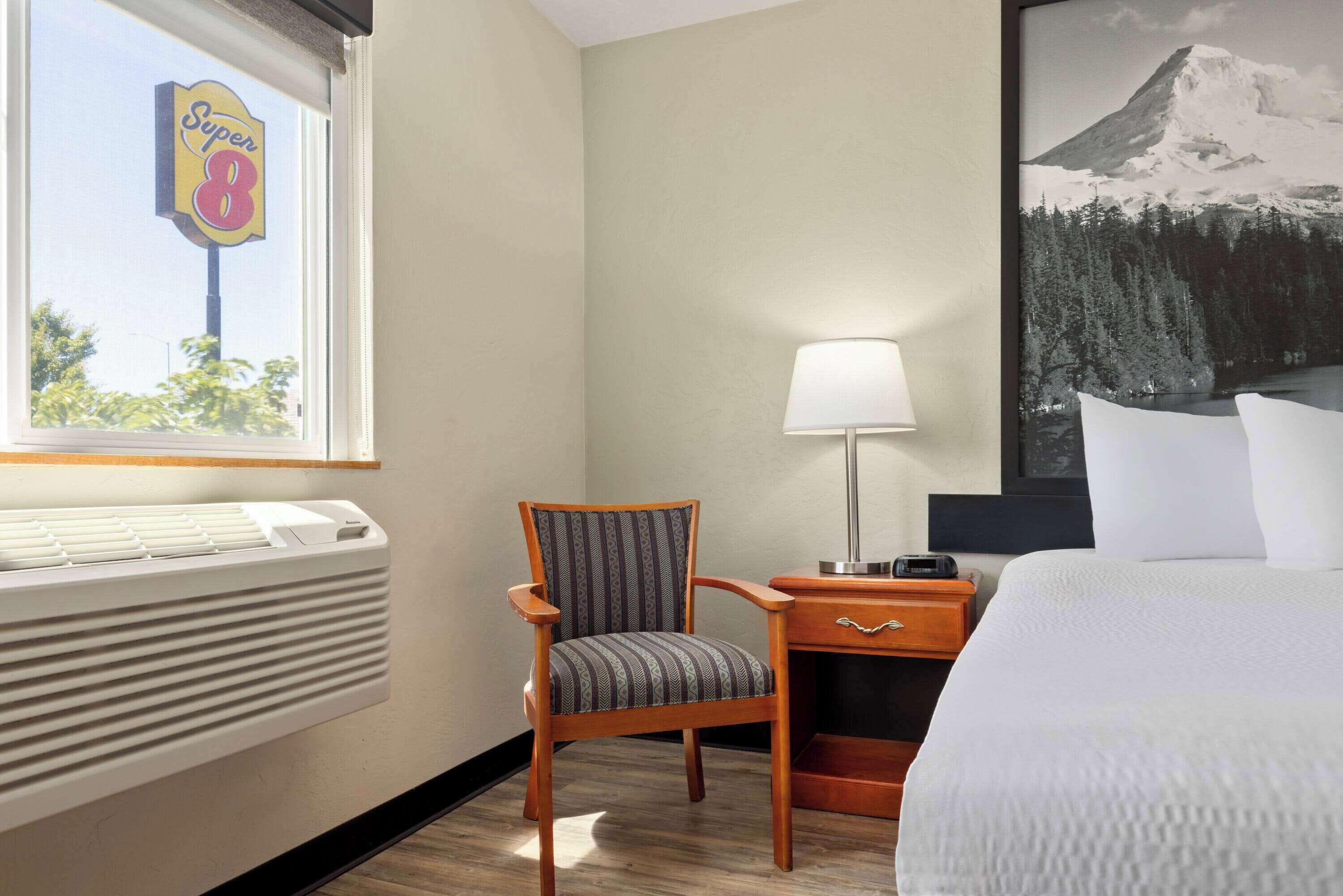 Une chambre d’hôtel avec un lit, une chaise, une table de chevet avec une lampe, un radiateur et une fenêtre avec une pancarte.