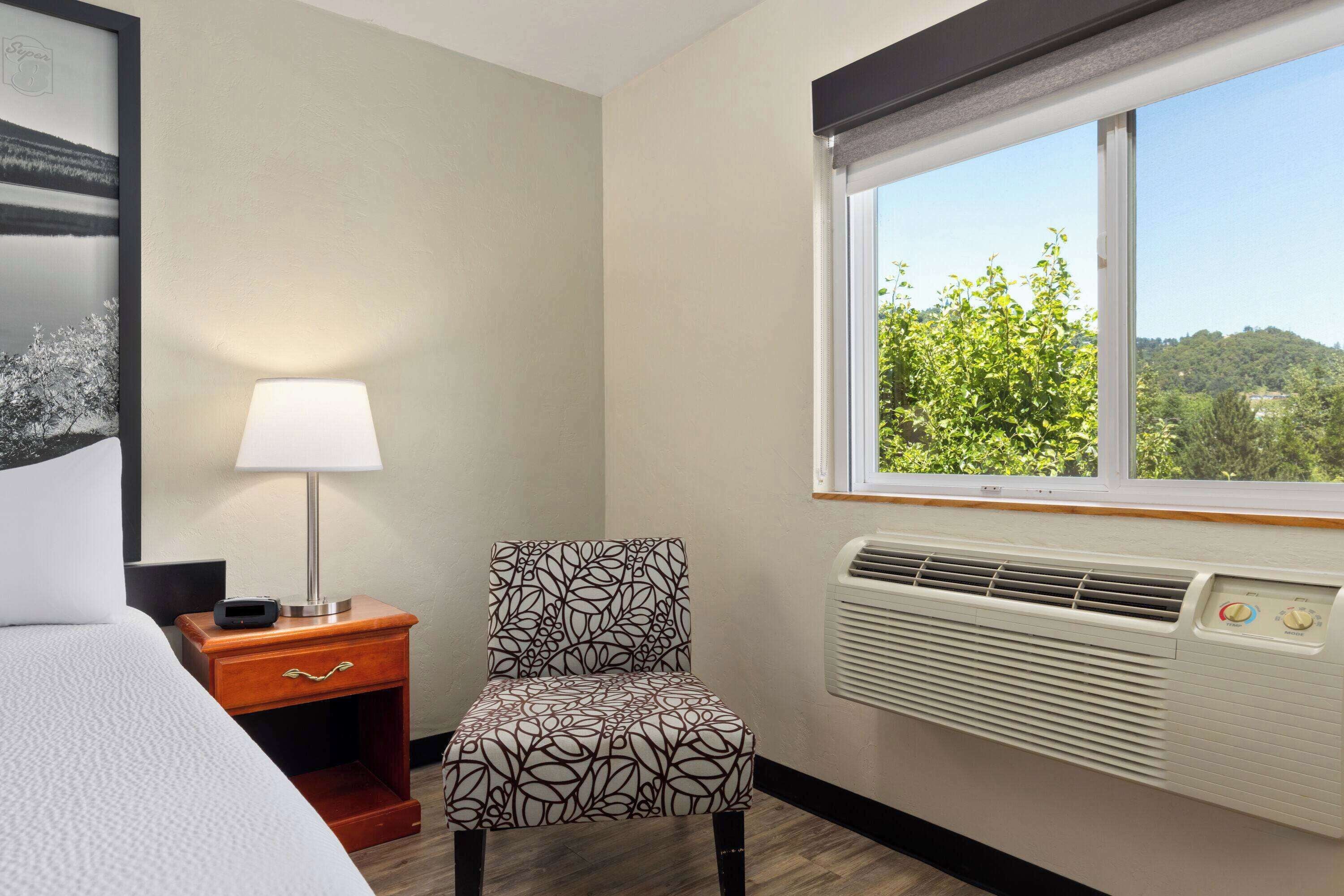 A hotel room with a bed, a nightstand with a lamp, a chair, a window with a view of greenery, and an air conditioning unit.