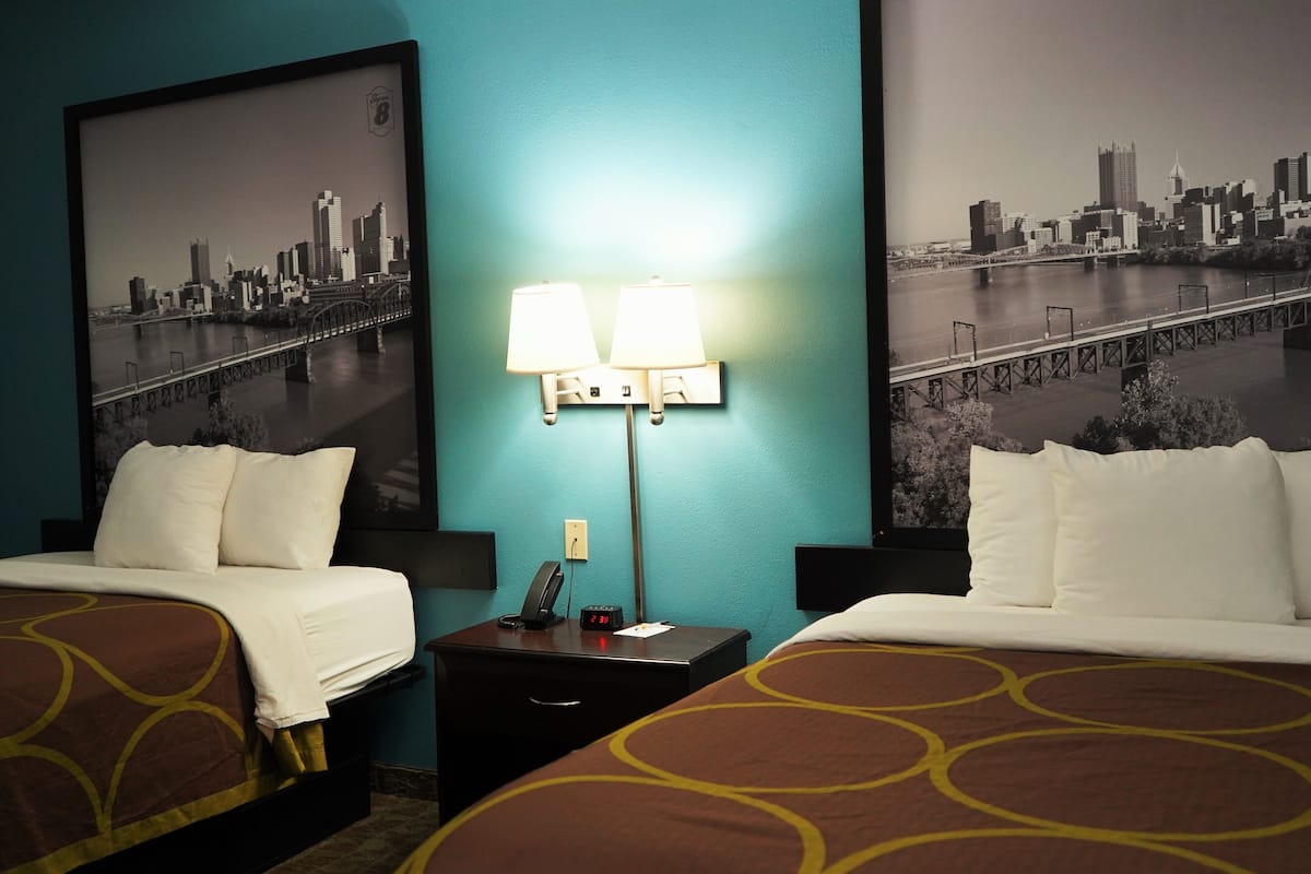 A hotel room with two beds, a nightstand with a lamp, and a cityscape view.