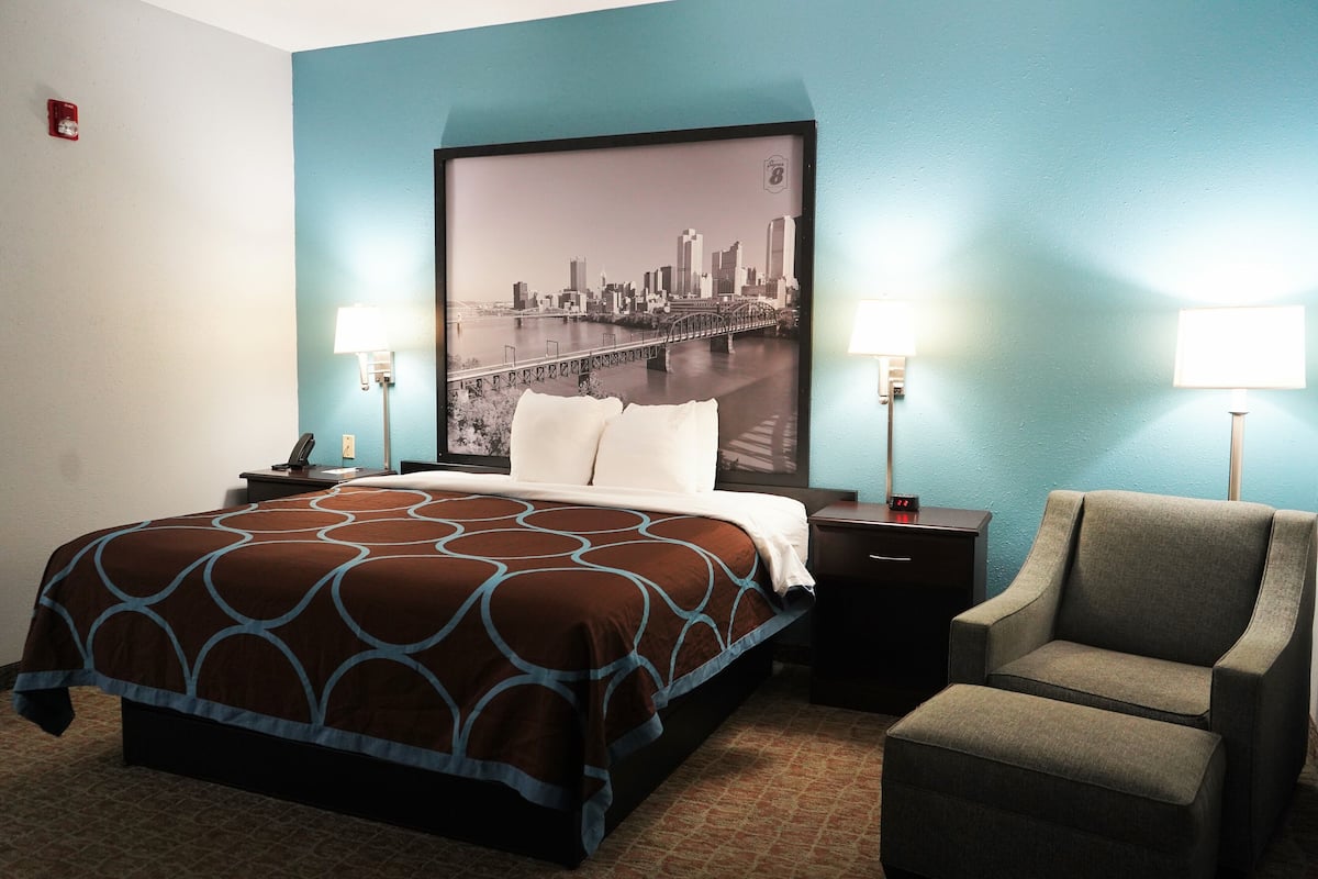 A hotel room with a bed, a chair, a nightstand, and a wall-mounted lamp.
