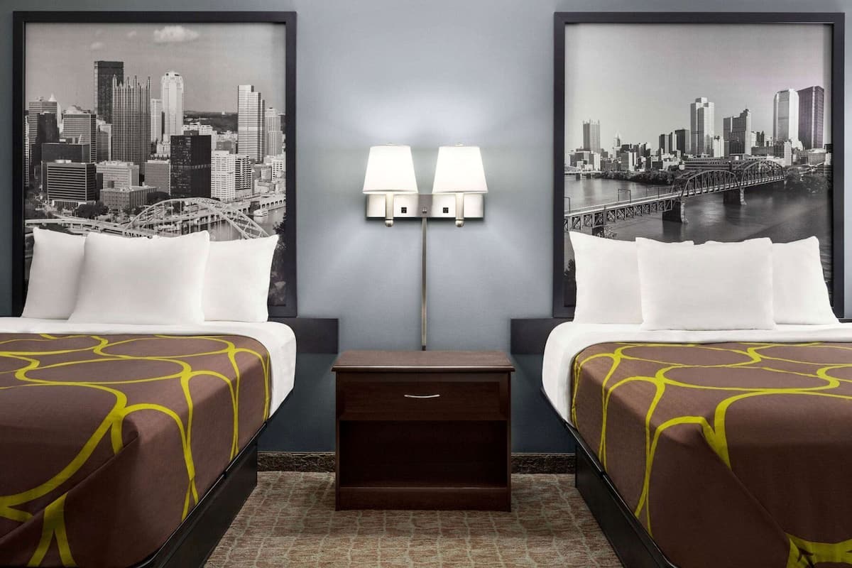 A hotel room with two beds, a nightstand, two wall-mounted lamps, and two large framed cityscape photographs.