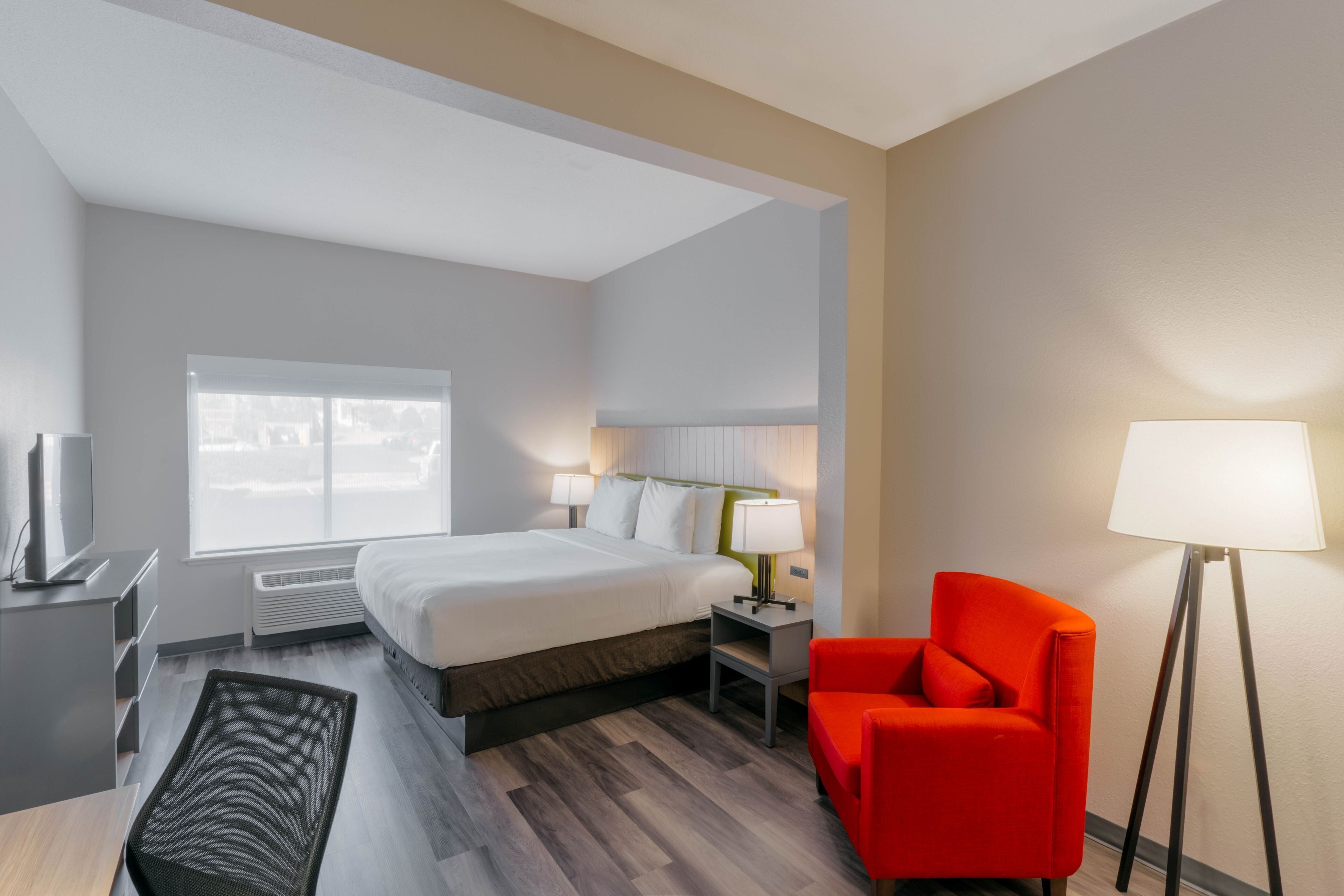 Quarto de hotel moderno com uma cama grande, luminárias de cabeceira, uma poltrona vermelha e uma escrivaninha com televisão de tela plana.