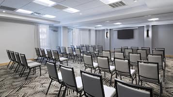 Une salle de conférence avec des rangées de chaises, une moquette à motifs et un téléviseur à écran plat accroché au mur.