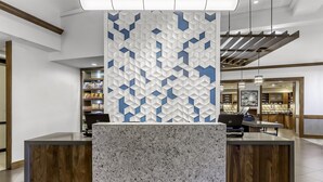 Zona de recepción con diseño moderno, destacando una pared con estampado geométrico, un mostrador de recepción de mármol y vistas al espacio contiguo con estanterías y mostrador.