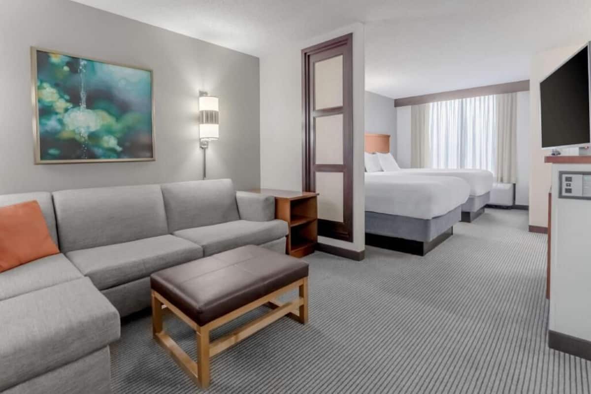 Una habitación de hotel con un sofá gris, una mesa de centro de madera, una cama con ropa blanca, un televisor y un cuadro en la pared.