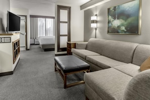 A hotel room with a sofa, a bench, a TV, a desk, and a painting. - Hyatt Place Charlotte Airport / Billy Graham Parkway (Charlotte)