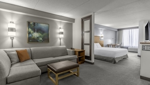 Una habitación de hotel con un sofá gris, un banco de madera, una cama, un escritorio y un cuadro en la pared.