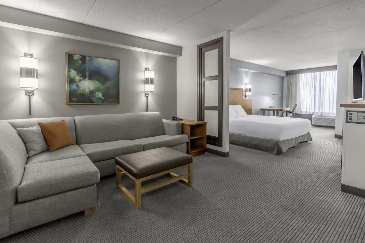 Una habitación de hotel con un sofá gris, un banco de madera, una cama, un escritorio y un cuadro en la pared.