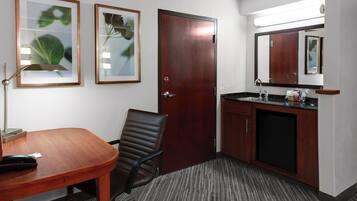 Habitación de hotel con escritorio de madera, una silla, un cuadro enmarcado, una puerta, un lavamanos y un espejo.