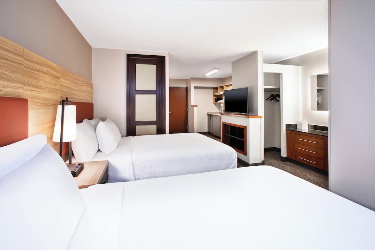 Ein Hotelzimmer mit zwei Betten, einem Schreibtisch mit Lampe, einem Fernseher und einer Küchenzeile.