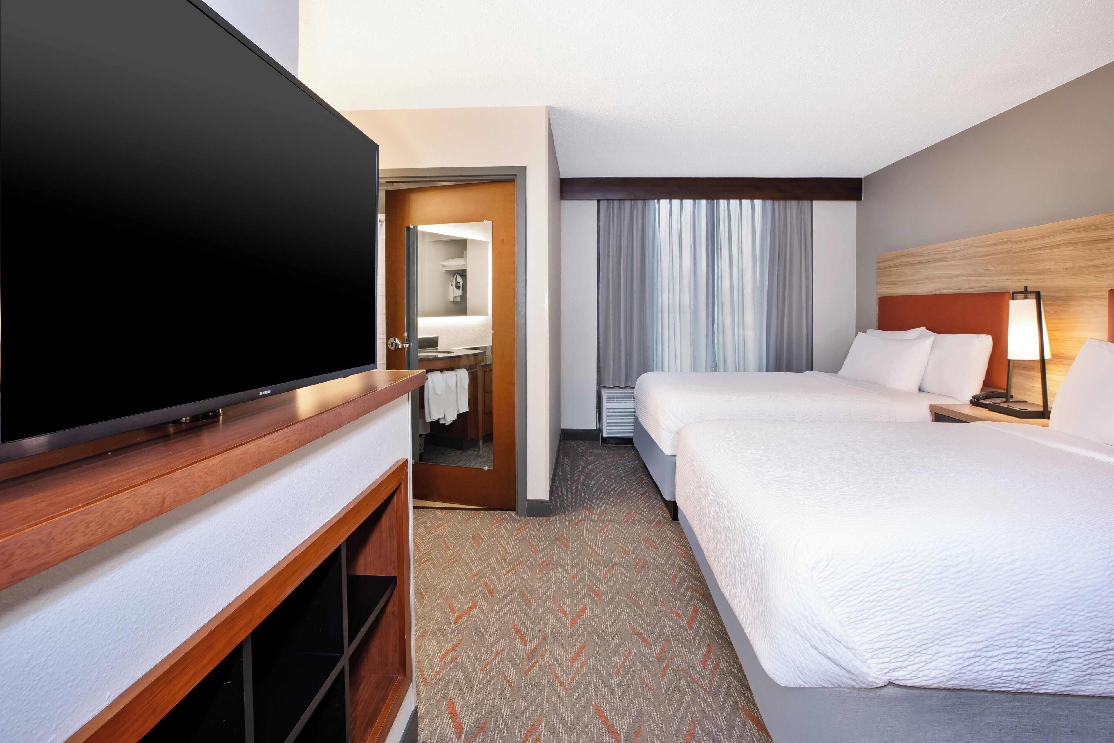 A hotel room with a flat-screen TV, two beds, a wooden TV stand, a mirror, a window with curtains, and a lamp.
