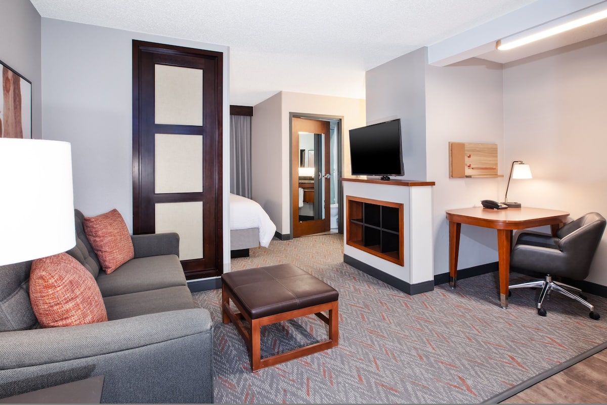 Ein Hotelzimmer mit einer Couch, einem kleinen Tisch, einem Fernseher, einem Schreibtisch mit Stuhl und einem durchgehenden Türspalt sichtbaren Badezimmer.