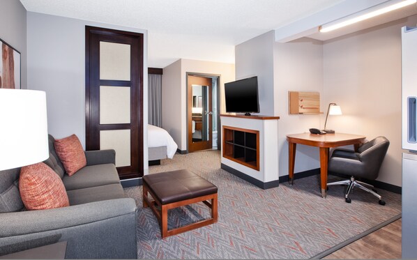 Habitación de hotel con sofá, una mesita, televisor, escritorio con silla y baño visible a través de una puerta abierta.